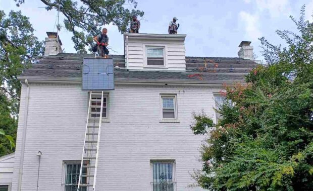 A solar installer pulls a solar panel up a ladder while two installers wait on the roof to bolt it in place.