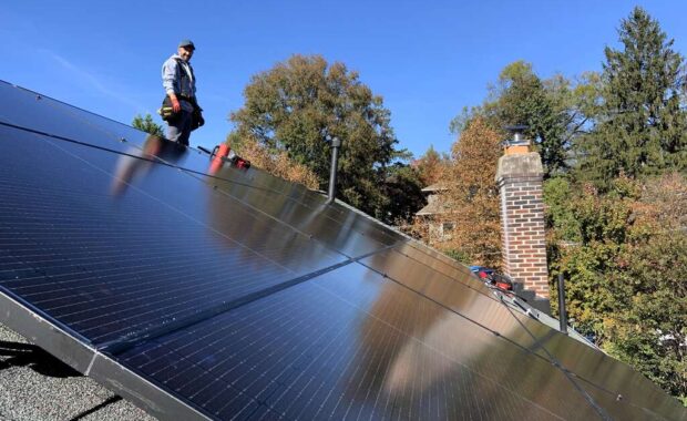 Black solar panels on a roof with a solar installer smiling in the background.