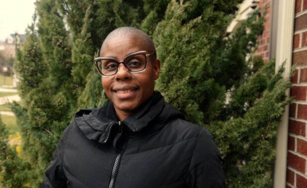 A woman wearing glasses and a black coat stands in front of greenery.